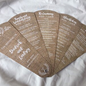 Puede incluir: Un abanico de boda de arpillera con texto blanco. El abanico incluye los detalles de la boda, la ceremonia, después del beso y el menú. El abanico está decorado con un diseño floral sencillo.