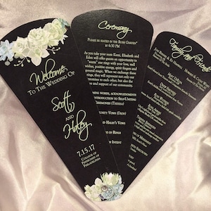 May include: Black and gold wedding program fan with a floral design. The program includes the ceremony order, the names of the wedding party, and the date of the wedding. The text reads "Welcome to the Wedding of Scott and Haley 7.15.17 College of Physicians of Philadelphia."