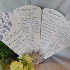 Könnte beinhalten: Hochzeitszeremonie-Programm-Fächer mit einem violetten Blumendesign und Schmetterlingen. Die Fächer enthalten die Details zur Hochzeitsgesellschaft, zur Zeremonie und zum Hochzeitsessen.