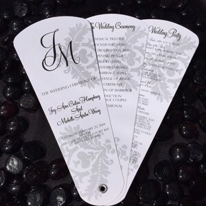 Puede incluir: Un abanico blanco de programa de ceremonia de boda con un diseño floral plateado. El programa incluye los nombres de la novia y el novio, Joy-Ann Calisa Humphrey y Michelle Andre Wray, la fecha de la boda y los detalles de la ceremonia. El abanico también incluye una lista de los invitados a la boda.