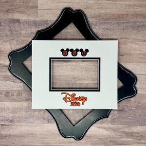 May include: A white picture frame mat with a black border, featuring the word "Boo" above the opening and the Disney logo with "2020" in orange. The mat is set on a black decorative frame.