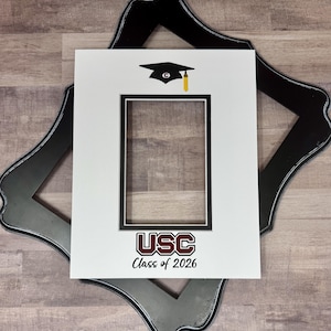 May include: A black and silver decorative frame with a white mat featuring a graduation cap graphic and the text "USC Class of 2026". The frame is designed to hold a photograph.