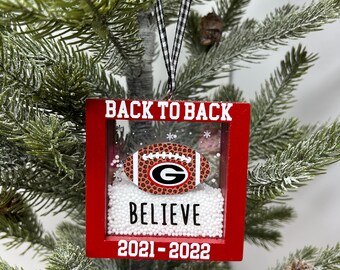 May include: Red and white Christmas ornament with a football, snow, and the text "Back to Back", "Believe", and "2021-2022".