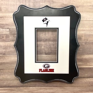 May include: Black and white picture frame with a white mat and a red and black logo that says "FLAGLINE" with a red "G" inside a circle. The logo is below a silhouette of a person holding a flag with a red "G" inside a circle.