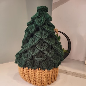 Puede incluir: Un gorro de árbol de Navidad de crochet verde y marrón. El gorro está hecho con un punto texturizado que se asemeja a escamas. La parte superior del gorro es puntiaguda y la parte inferior es redondeada.