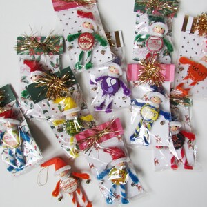 May include: A collection of ten handmade elf ornaments packaged in clear plastic sleeves. Each elf features a bottle cap torso, pipe cleaner limbs in various colors, a small painted wooden head, a felt hat, and gold tinsel accents.