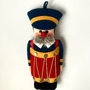 May include: A plush toy soldier with a blue hat and uniform, accented with yellow trim and red straps. The soldier has a red nose, gray beard, and a red drum with gold accents. A loop is attached to the top of the hat.