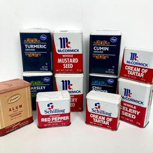 May include: A collection of vintage spice tins, including McCormick and Schilling brands. The tins feature labels with text such as "Turmeric Ground," "Mustard Seed," and "Cream of Tartar." The tins are in various colors, including red, white, and navy blue.