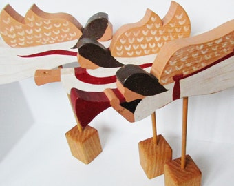 May include: Three wooden angel figurines with painted details. Each angel has a white and brown wing, a red and white striped dress, and a brown face. The angels are standing on wooden bases.