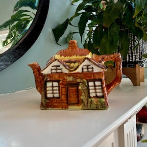 May include: A ceramic teapot shaped like a cottage, featuring a brown thatched roof, white-framed windows, and a brown door. The teapot is decorated with green vines and sits on a white surface. The background includes a mirror and a potted plant.