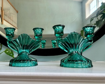 May include: Two teal glass candle holders with a seashell design. Each holder has three candle cups. The holders are on a white surface, with a large round mirror in the background.