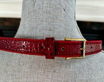 May include: A close-up of a red belt with a textured, crocodile-like pattern. The belt has a gold-colored rectangular buckle and a matching belt loop. The belt is displayed on a mannequin.