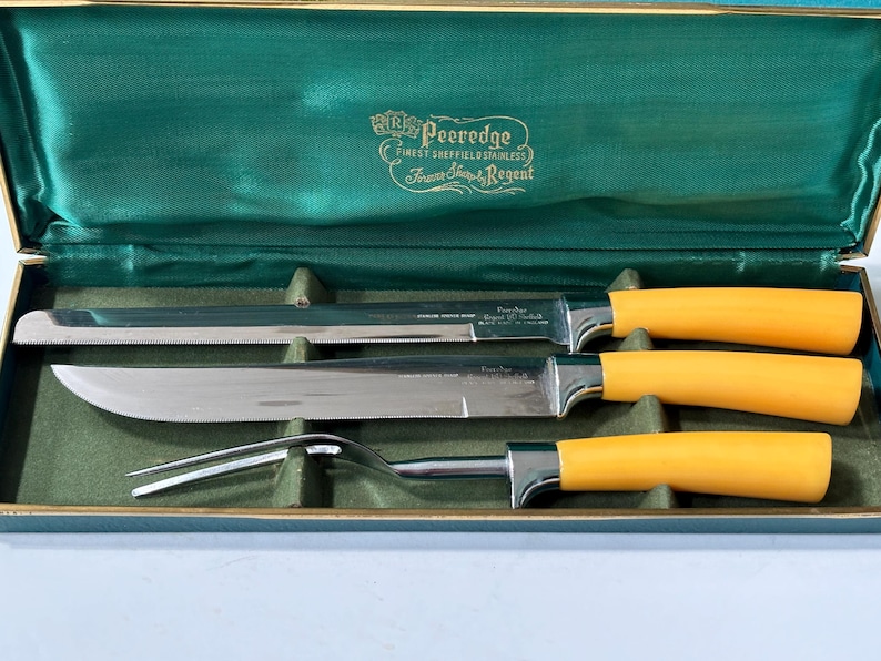 K&ouml;nnte beinhalten: Ein Vintage-Besteckset in einem gr&uuml;n mit Samt ausgekleideten Etui. Das Set enth&auml;lt ein Tranchiermesser, ein Aufschnittmesser und eine Tranchiergabel, alle mit Klingen aus Edelstahl und gelben Griffen. Das Etui ist mit dem Markennamen "Peeredge" gepr&auml;gt.