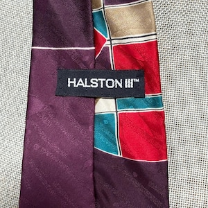 May include: A necktie featuring a deep purple fabric with a subtle diagonal textured pattern. A black label with the white text HALSTON III is stitched across the center. The right side displays a geometric grid pattern in red, teal, and gold.