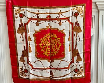 May include: A red, white, and gold silk scarf with a decorative design. The scarf features a red border, a white center with gold rope and tassel details, and a central red emblem with gold lettering.