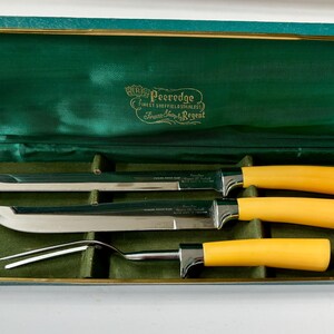 K&ouml;nnte beinhalten: Ein Vintage-Tranchierbesteck in einer gr&uuml;n samtgef&uuml;tterten Box. Das Set enth&auml;lt ein Tranchiermesser, ein Aufschnittmesser und eine Tranchiergabel. Die Griffe sind leuchtend gelb und die Klingen aus Edelstahl. Die Box hat goldfarbene Verzierungen und den Text "Peeledge".