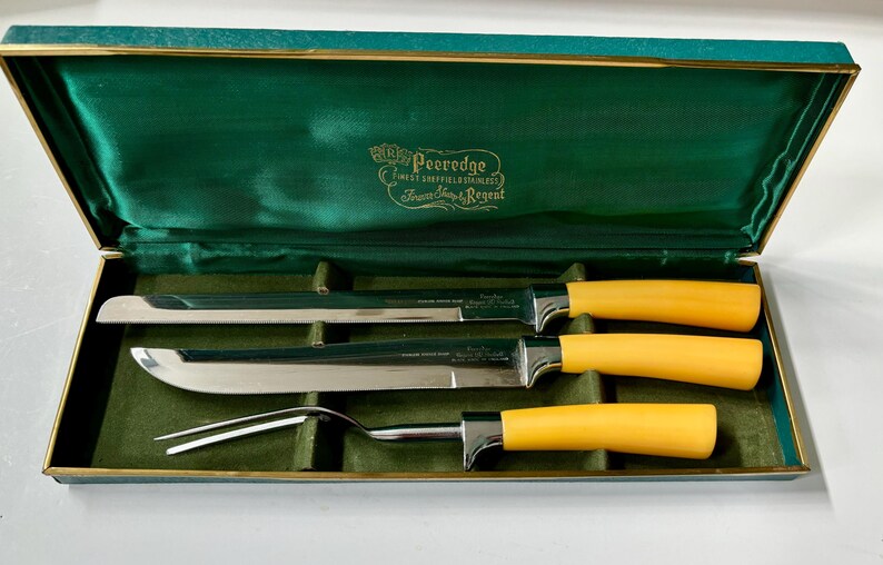 K&ouml;nnte beinhalten: Ein Vintage-Tranchierbesteck in einer gr&uuml;n samtgef&uuml;tterten Box. Das Set enth&auml;lt ein Tranchiermesser, ein Aufschnittmesser und eine Tranchiergabel. Die Griffe sind leuchtend gelb und die Klingen aus Edelstahl. Die Box hat goldfarbene Verzierungen und den Text "Peeledge".