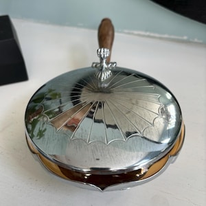 May include: A silver-colored, round, lidded dish with a wooden handle. The lid has a sunburst design. The dish has a scalloped edge and a decorative finial where the handle attaches. The interior appears to be gold-toned.