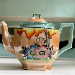 May include: A vintage teapot with a peach-colored body, adorned with a floral design in blue, pink, and green. The lid and handle are mint green, and the spout is peach. The teapot has a square handle and a matching lid with a small square knob.