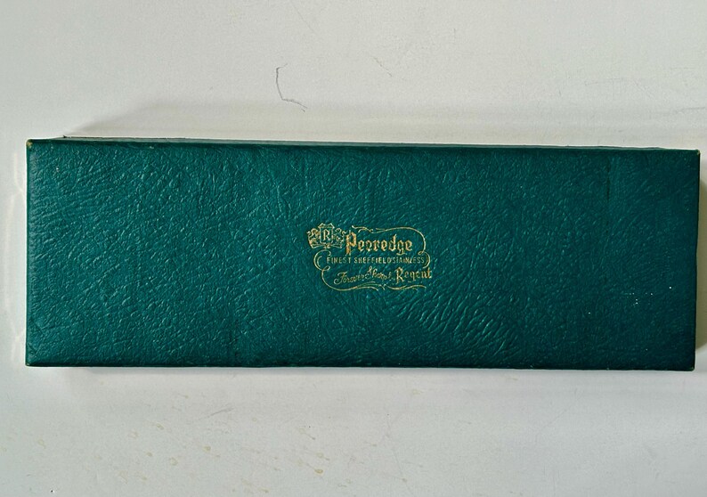 K&ouml;nnte beinhalten: Eine rechteckige, teal-farbene Box mit strukturierter Oberfl&auml;che. Auf der Box steht in goldener Schrift "Pezredge, Finest Sheffield Cutlery, Trafalgar House, Regent."
