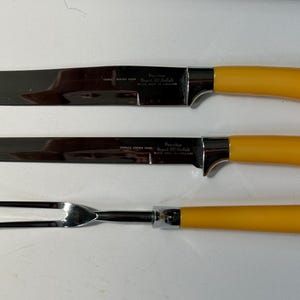 K&ouml;nnte beinhalten: Ein dreiteiliges Tranchier-Set mit Klingen aus Edelstahl und hellgelben Griffen. Das Set enth&auml;lt ein Tranchiermesser, ein Aufschnittmesser und eine Tranchiergabel. Die Klingen sind leicht gebogen und mit Text versehen. Die Griffe sind einfarbig.
