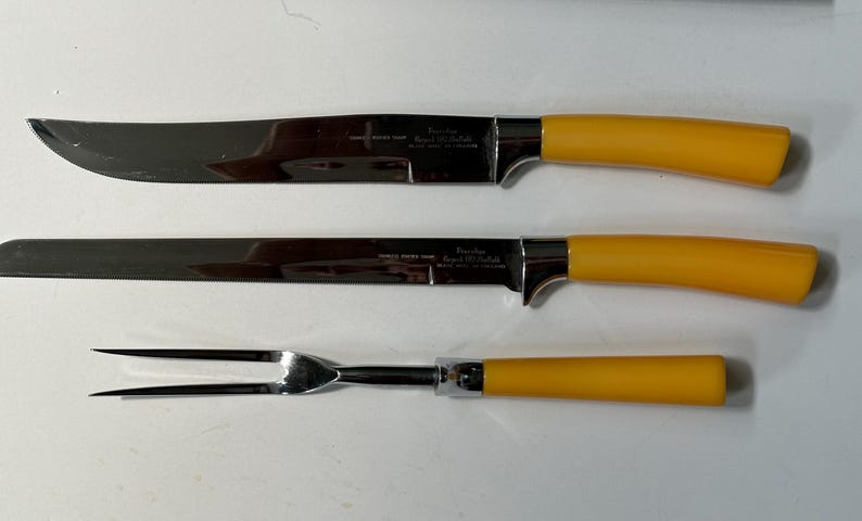 K&ouml;nnte beinhalten: Ein dreiteiliges Tranchier-Set mit Klingen aus Edelstahl und hellgelben Griffen. Das Set enth&auml;lt ein Tranchiermesser, ein Aufschnittmesser und eine Tranchiergabel. Die Klingen sind leicht gebogen und mit Text versehen. Die Griffe sind einfarbig.