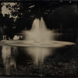 May include: A black and white photograph of a fountain in a pond surrounded by trees. The fountain is spraying water high into the air, creating a misty effect.