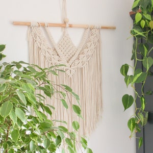 May include: A white macrame wall hanging with a wooden dowel rod at the top. The macrame is in a triangular shape with a fringe at the bottom.