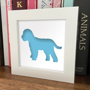 May include: A white frame with a light blue silhouette of a dog inside. The frame is sitting on a wooden surface.