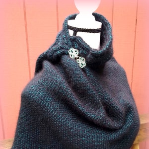 May include: A black and teal knitted shawl with a decorative silver clasp. The shawl is draped over a white mannequin.