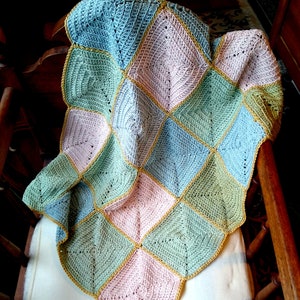 Pode incluir: Um cobertor de bebé de crochê com um design de patchwork em tons de azul, verde e rosa. O cobertor é guarnecido com um fio dourado.