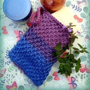 May include: A purple and blue crocheted soap saver with a loop handle. The soap saver is on a green background with a white bar of soap, a blue jar, and a copper-colored sphere.