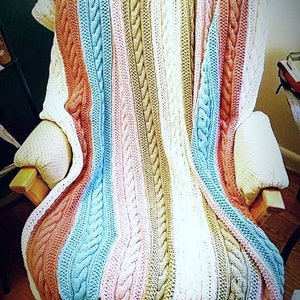 May include: A striped knitted throw blanket with a cable knit pattern in shades of pink, blue, brown, and white. The blanket is draped over a wooden chair.