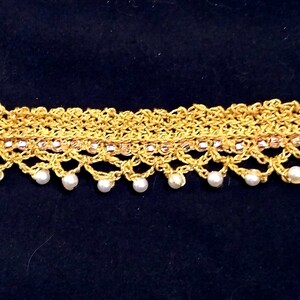 Peut inclure: Un collier ras du cou en crochet doré au design délicat. Le collier présente une rangée de petites perles blanches nacrées suspendues au bord inférieur. Une rangée de petites perles roses est également visible.