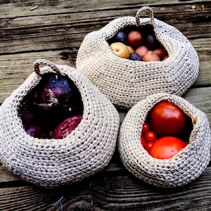 May include: Three crocheted baskets filled with produce. The first basket is filled with red onions, the second with red and purple potatoes, and the third with red tomatoes.