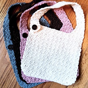 Puede incluir: Cuatro baberos de ganchillo en diferentes colores: blanco, rosa, marrón y azul. Cada babero tiene un cierre de botón y una textura de ganchillo.