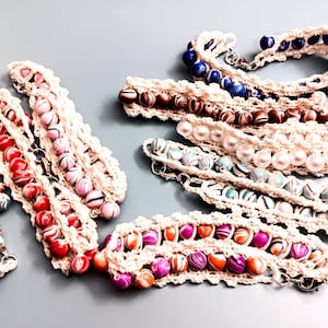 May include: Several crocheted necklaces with colorful beads. The beads vary in color, including red, pink, blue, and white. The crochet work is a cream color, and the necklaces are arranged on a light gray surface.