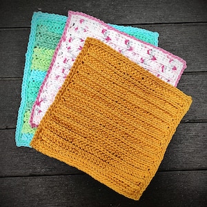 May include: Three crocheted dishcloths in different colors and patterns. The top dishcloth is pink and white with a floral pattern. The middle dishcloth is light blue and green with a striped pattern. The bottom dishcloth is a solid mustard yellow.