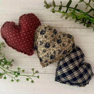 May include: Three heart-shaped fabric ornaments. One is burgundy with white dots, another is beige with blue floral patterns, and the third is navy blue plaid. Each has a frayed edge.