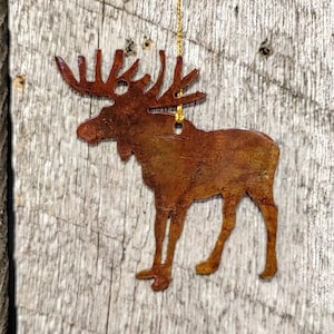 May include: A rusty brown metal moose ornament with a gold chain for hanging. The moose is standing on a weathered wooden surface.