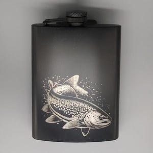 Pode incluir: Frasco preto de aço inoxidável com uma ilustração gravada de um peixe. O frasco tem uma tampa de rosca e formato retangular. O desenho do peixe é branco sobre fundo preto. Um acessório elegante para os amantes da natureza.