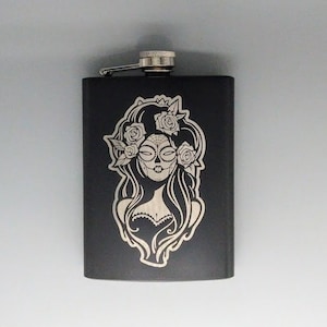 May include: Black stainless steel flask with a laser engraved design of a woman with a sugar skull face and flowers in her hair.