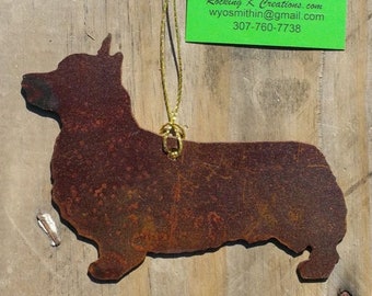 May include: A rusty brown metal ornament shaped like a corgi dog. The ornament is hanging from a gold string. The ornament is on a wooden background. The text on the tag reads "Rocking K Creations.com wyosmithin@gmail.com 307-760-7738"