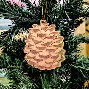 Puede incluir: Un adorno de Navidad de madera con forma de piña. El adorno es marrón y tiene un diseño detallado. Está colgado de una cuerda dorada en un árbol de Navidad artificial verde.