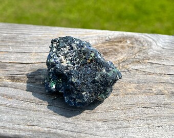 May include: A dark blue and green rough mineral rock with white flecks. The rock is sitting on a weathered wooden surface.