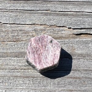 May include: A pink and white rough cut gemstone with a hexagonal shape. The gemstone is sitting on a weathered wooden surface.