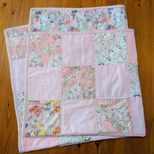 May include: A patchwork quilt in shades of pink and floral patterns. The quilt is made up of squares of solid pink, white polka dots, and floral prints, with a pink and white striped border. The quilt is folded on a wooden surface.