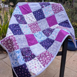 May include: A patchwork quilt with a variety of floral patterns in shades of purple, blue, and white. The quilt is made up of squares and is folded in half.