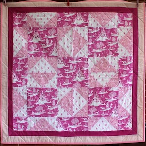 May include: A pink and white patchwork quilt with a repeating pattern of a castle, trees, and animals. The quilt is made of squares and triangles and has a pink border.