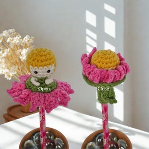 Op de afbeelding: Twee gehaakte bloemvormige potloodtoppers. De linker topper, gelabeld Open, toont een klein figuurtje met geel haar op een groene basis met roze bloemblaadjes. De rechter topper, gelabeld Close, heeft een geel centrum omringd door roze blaadjes.